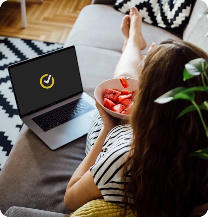 Mujer tumbada en un sofá comiendo un bol de fruta con un portátil a su lado con el logotipo de Norton